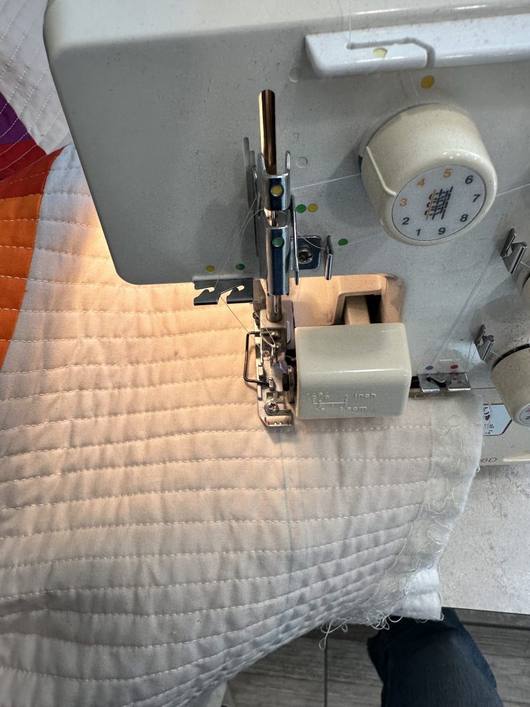 A close-up view of a sewing machine stitching through quilted fabric, with the machine's needle and presser foot clearly visible.