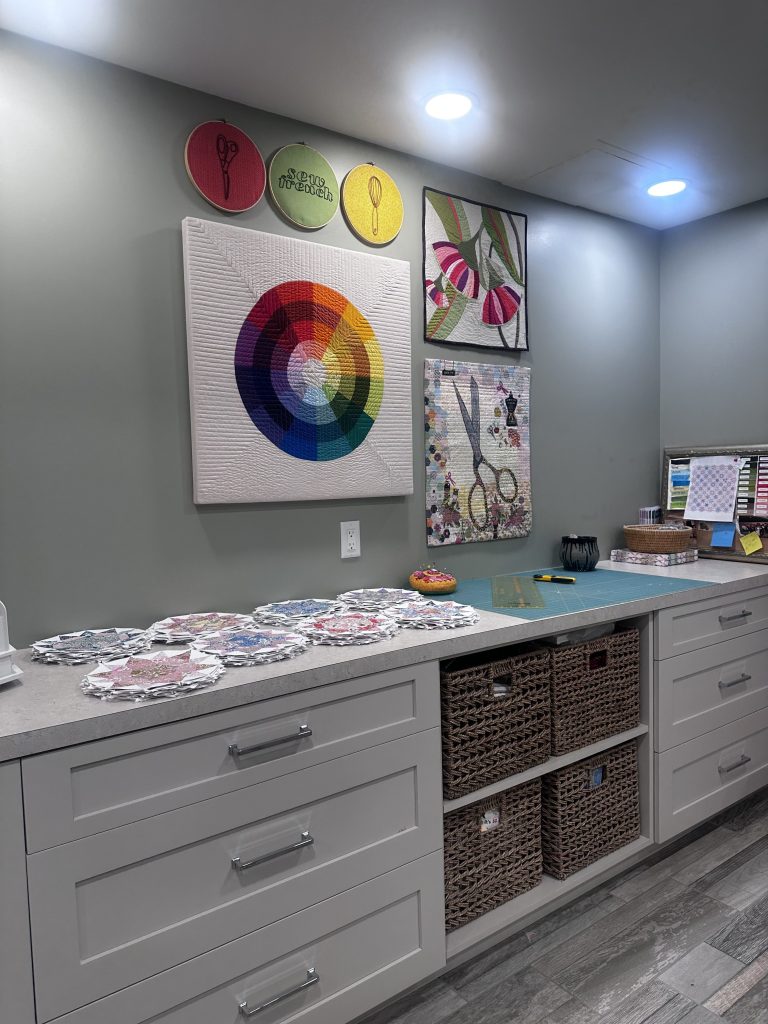 A creative workspace featuring a color wheel quilt on the wall, along with decorative circle signs and colorful art pieces. A countertop displays various fabric pieces, while storage drawers and baskets are organized below.