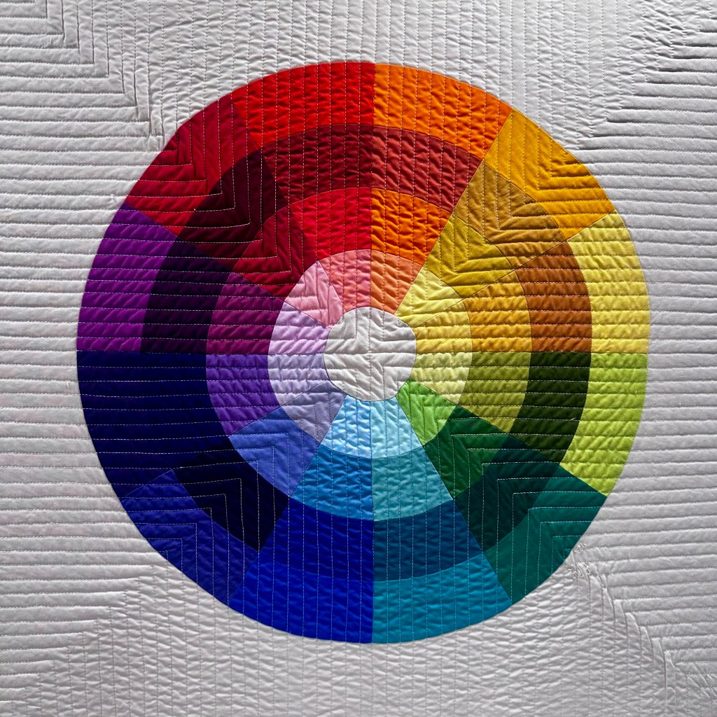 Color wheel quilt featuring a vibrant arrangement of colors in circular segments on a textured white background.