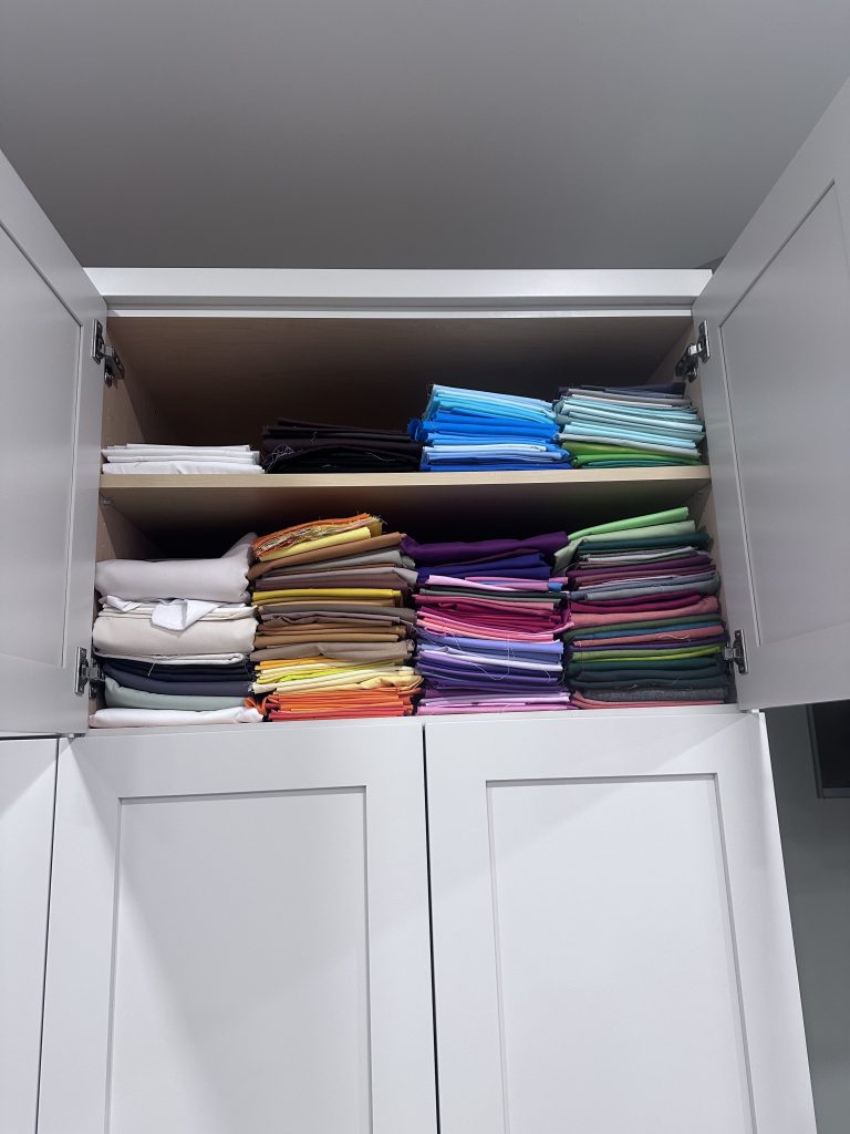 A neatly organized shelf in a cabinet displaying folded fabric in various colors, including blue, purple, green, yellow, pink, and white.