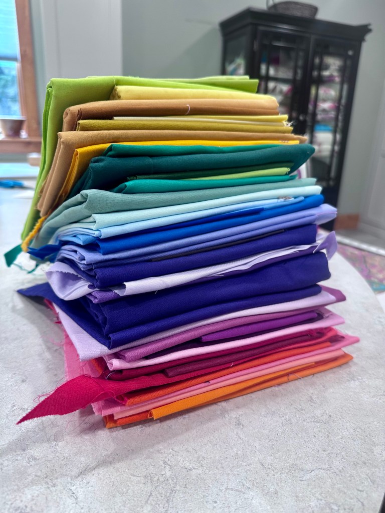 A colorful stack of folded fabric in various shades, including green, yellow, blue, purple, pink, and orange, arranged neatly on a light surface.