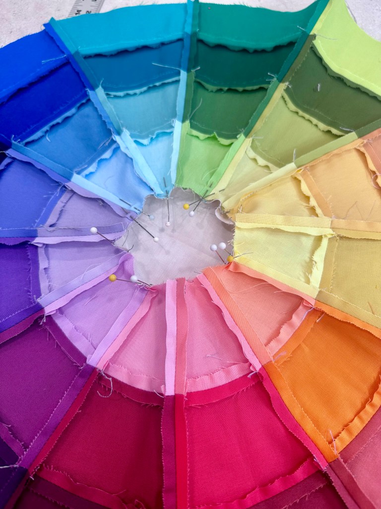 A circular arrangement of fabric swatches in various colors, pinned together at the center. The colors range from blue, green, yellow, orange, to purple, creating a gradient effect.