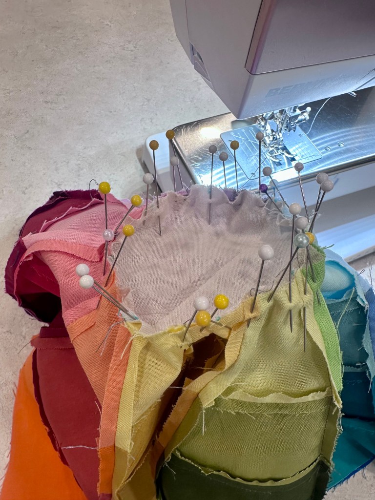 A close-up view of a sewing machine with colorful fabric pieces pinned together, showing various sewing pins secured around the edges.