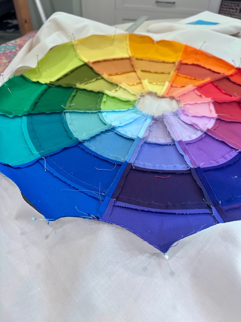 A vibrant fabric color wheel composed of layered fabric pieces in various shades, including red, orange, yellow, green, blue, and purple, pinned onto a white surface.