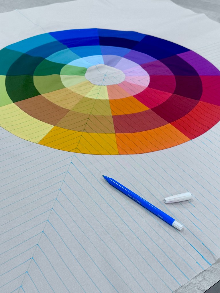 A color wheel illustration featuring various color segments arranged in a circular pattern, placed on a lined paper surface with a blue pen and a white eraser nearby.