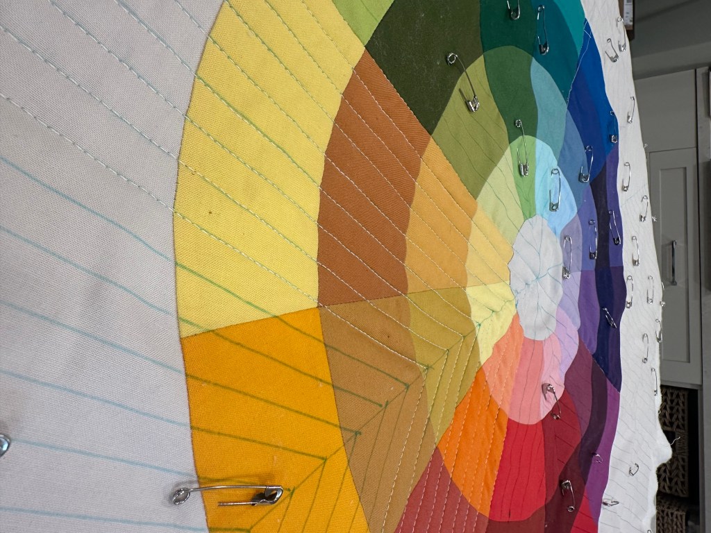 A close-up of a colorful fabric color wheel, showcasing various shades sewn into a quilted pattern with safety pins holding sections together.