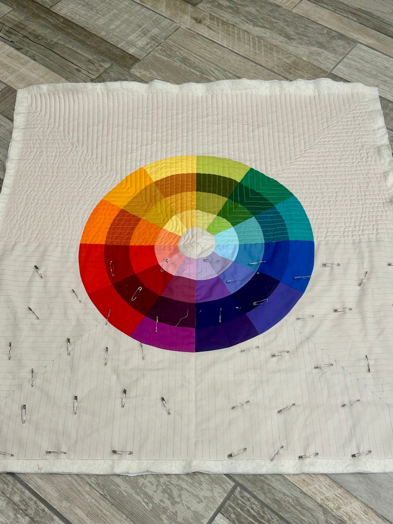 A colorful quilt featuring a circular gradient color wheel in the center, surrounded by white fabric with safety pins attached for quilting preparation.