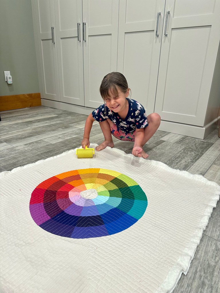 A smiling child is crouching on the floor, using a yellow roller to paint a colorful circular pattern on a white quilted fabric. The design features a spectrum of colors arranged in a circular layout.