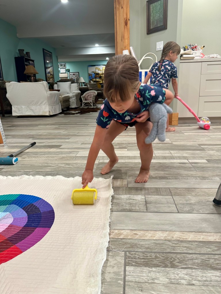 A young child is sitting on the floor in a living room, rolling a small yellow paint roller over a colorful blanket with a circular design. The child is holding a stuffed animal in one arm and focused on the task.